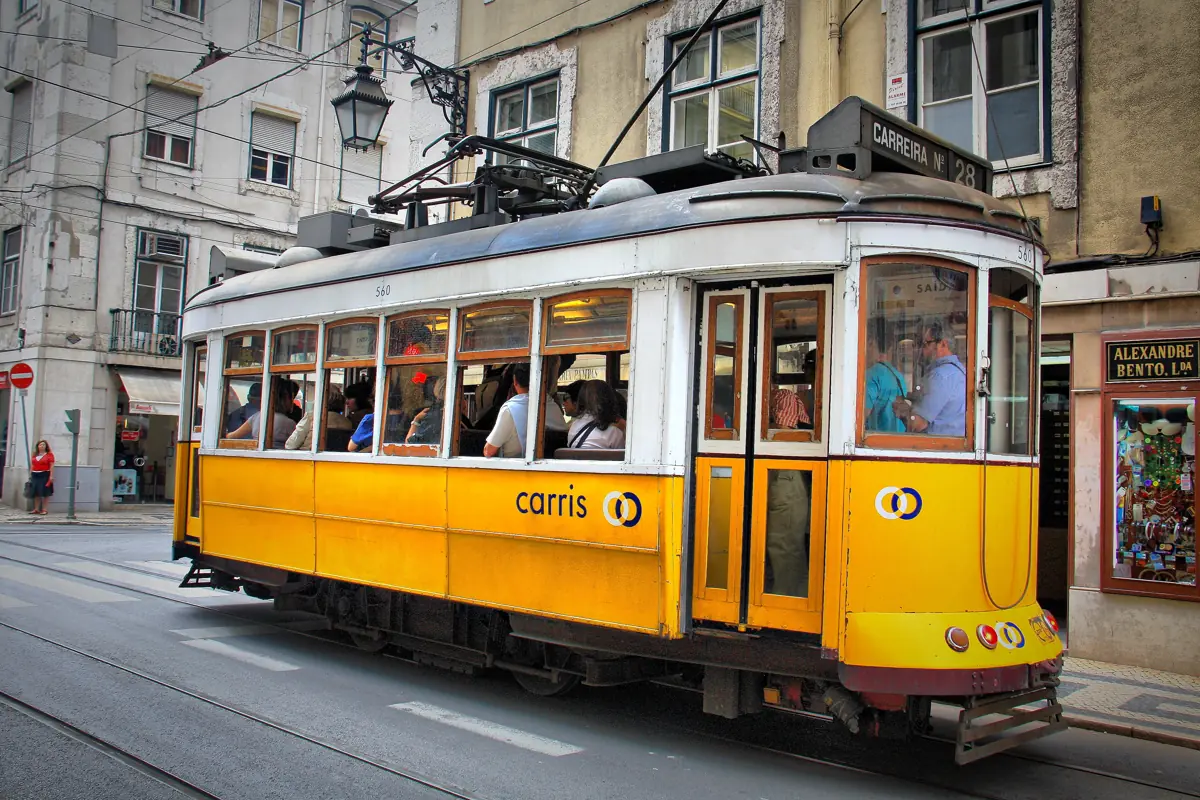 A yellow Carris number 28 tram full of passengers passing a shopfront on a narrow Lisbon street